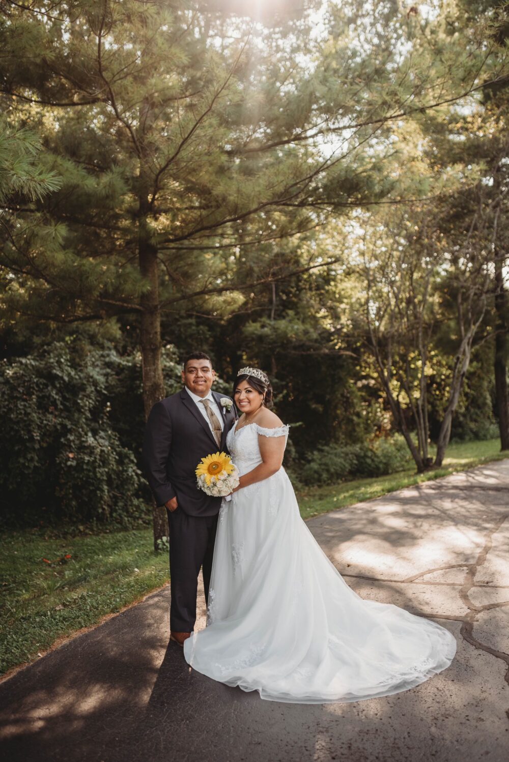 A bride and groom stand together on a sunlit path surrounded by trees, with the bride wearing an off-the-shoulder gown and tiara while holding a sunflower bouquet.