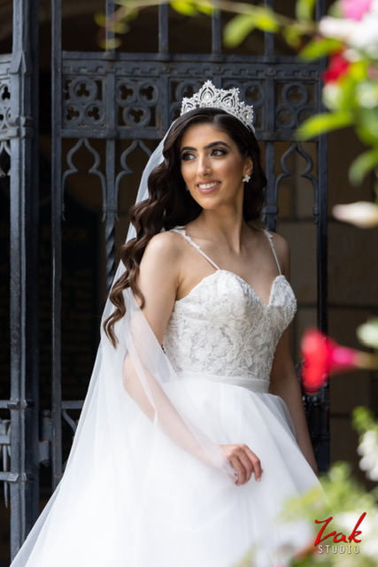 A bride wearing a tiara and veil stands near an ornate gate in a lace bodice wedding dress, looking off to the side with a soft smile.