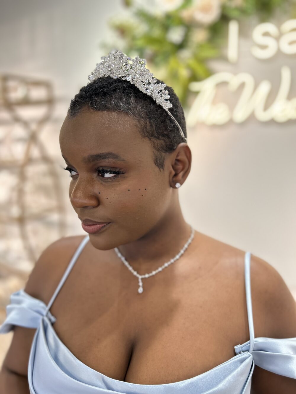 A woman with short hair wears a crystal tiara and necklace, posing in a soft blue dress against a blurred indoor background.