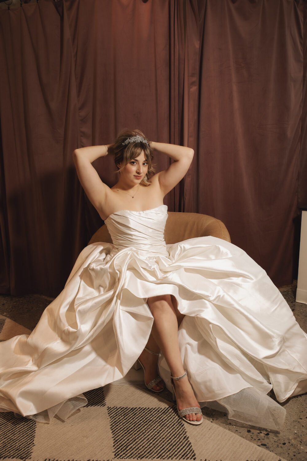 A bride sits in a chair wearing a strapless satin wedding dress and tiara, posing in front of brown draped curtains.