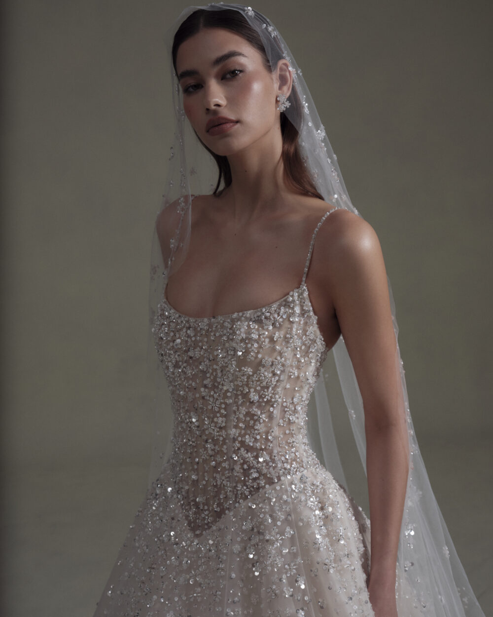 Close-up portrait of a model wearing a beaded sheer bodice wedding gown and embellished veil against a dark backdrop.