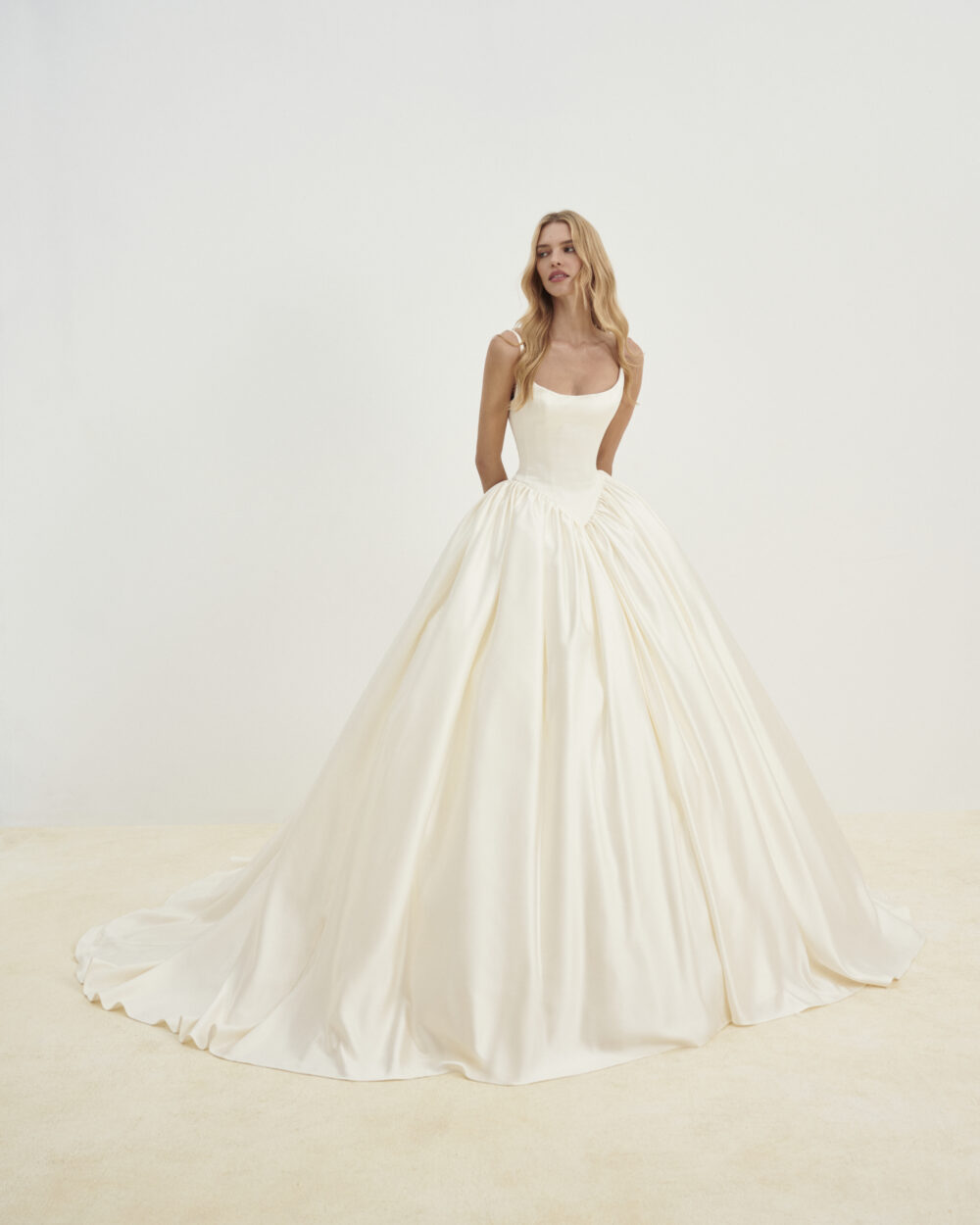 Model wearing a satin ball gown wedding dress with thin straps and a full skirt posed against a light studio background.