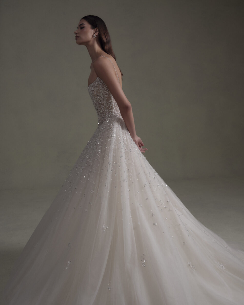 Model in a sparkling sleeveless ball gown wedding dress shown in side profile against a dark neutral backdrop.