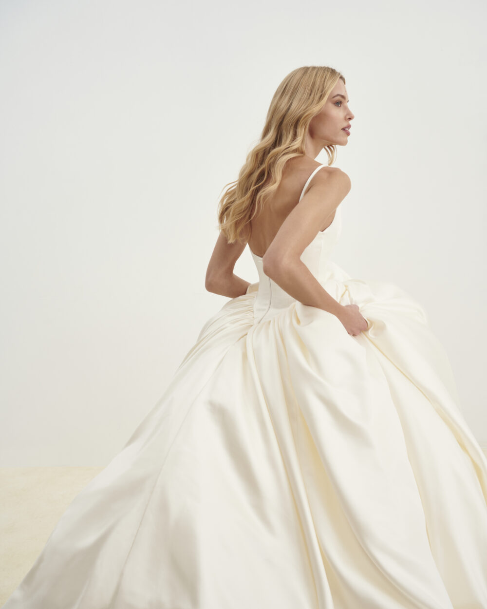 Model in an ivory satin ball gown wedding dress viewed from the back while holding the skirt in a studio setting.
