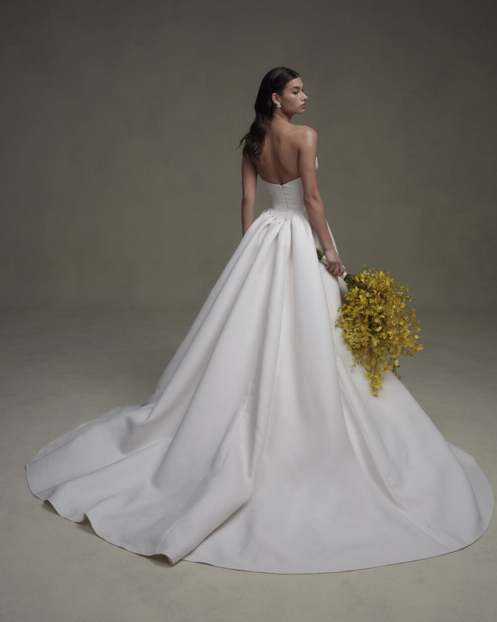 Model shown from the back in a strapless wedding gown with a full satin overskirt while holding a yellow flower bouquet.