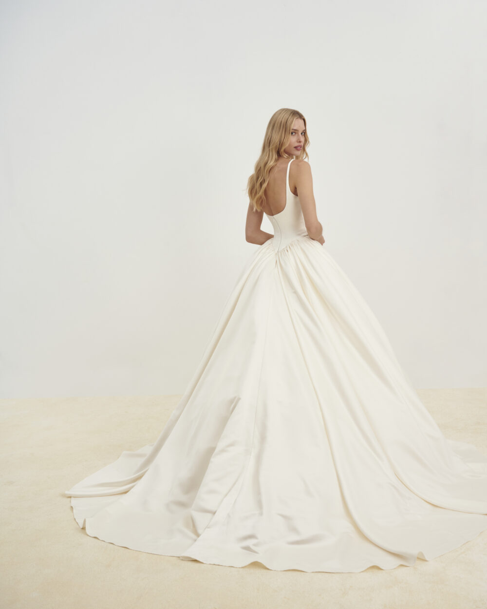 Model shown from the back in an ivory satin ball gown wedding dress with thin straps and a sweeping train in a studio.