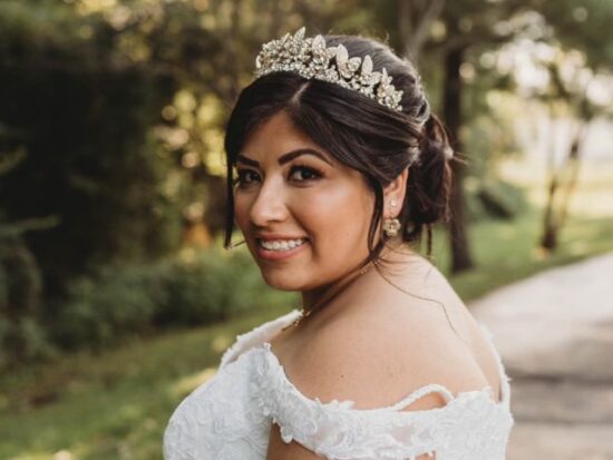 A bride wearing a tiara and lace gown smiles over her shoulder outdoors, holding a bouquet with a large sunflower.