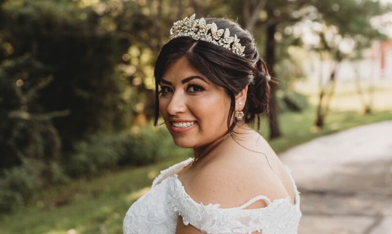 A bride wearing a tiara and lace gown smiles over her shoulder outdoors, holding a bouquet with a large sunflower.