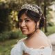 A bride wearing a tiara and lace gown smiles over her shoulder outdoors, holding a bouquet with a large sunflower.
