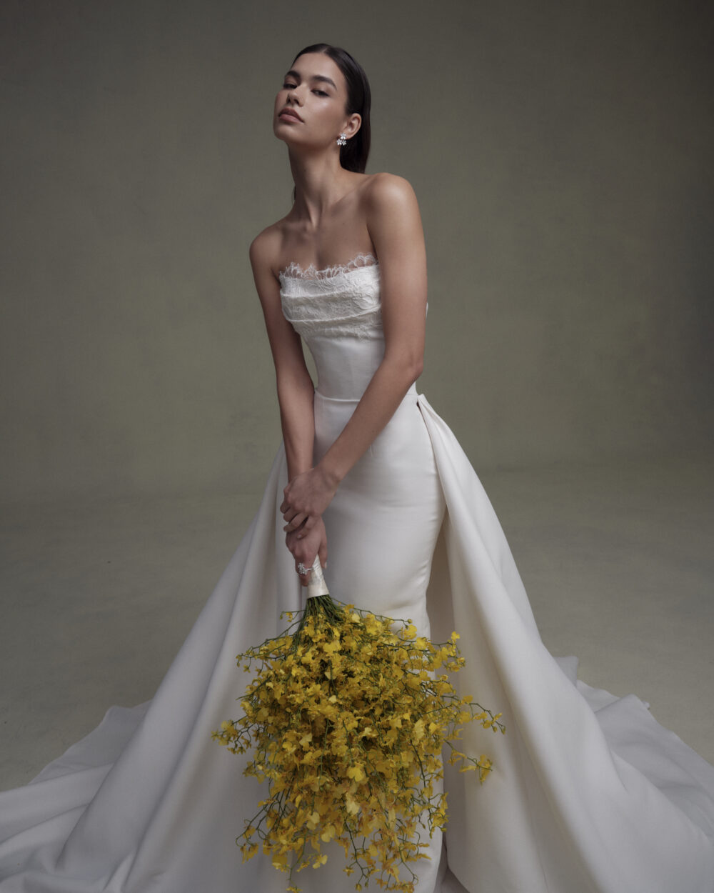 Model in a fitted strapless wedding gown with overskirt holds a cascading bouquet of yellow flowers against a dark backdrop.