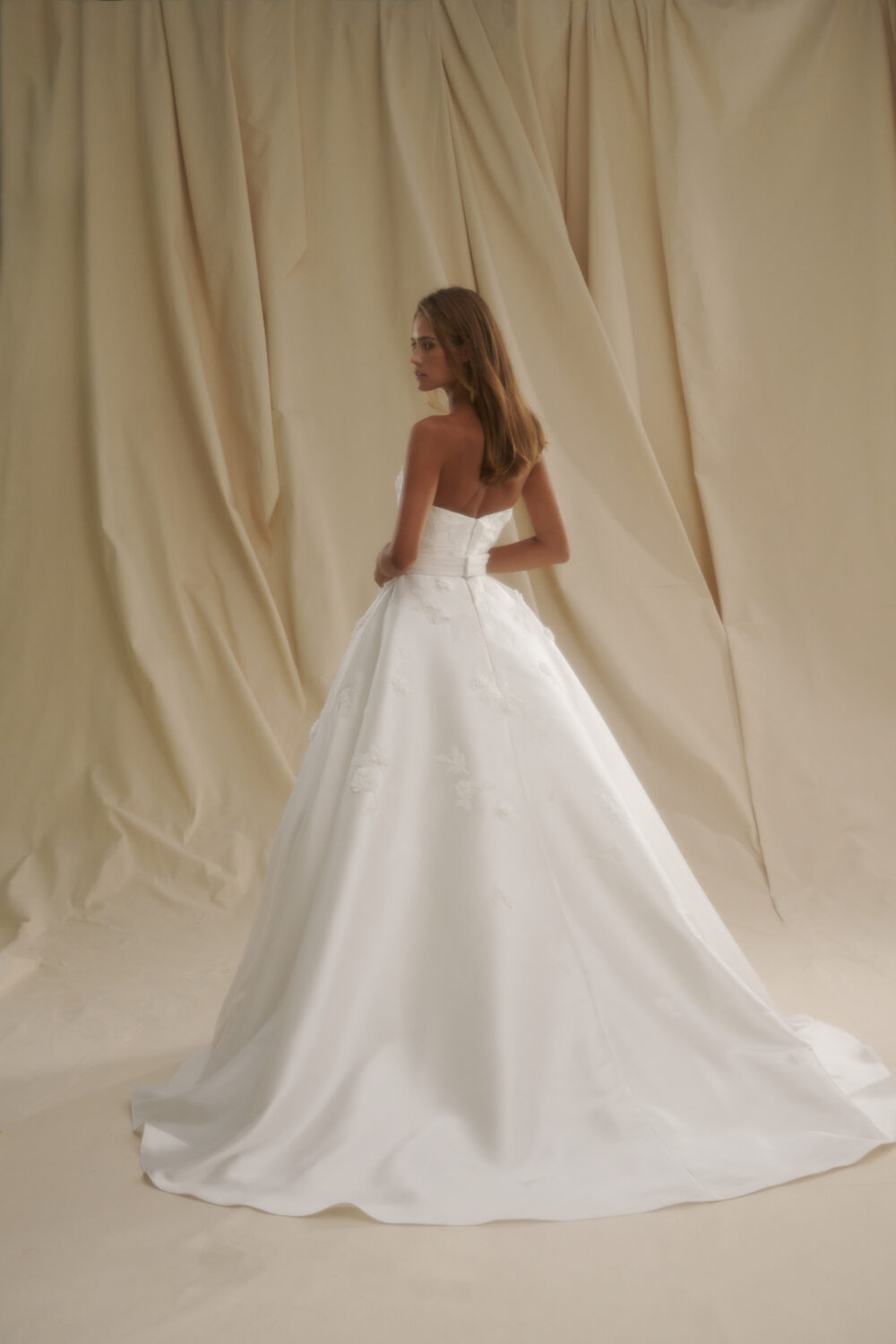 Model shown from the back in a strapless satin ball gown wedding dress with floral accents against draped fabric panels.