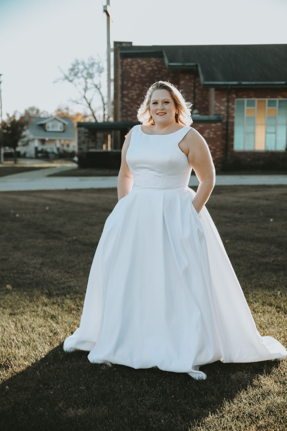 Bride in a sleeveless satin ball gown stands on a grassy lawn with a brick church building in the background.