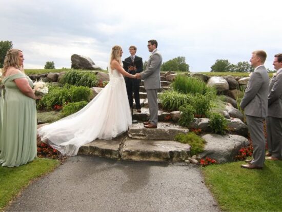 Outdoor wedding ceremony with the bride and groom exchanging vows on a stone platform, surrounded by bridesmaids and groomsmen.