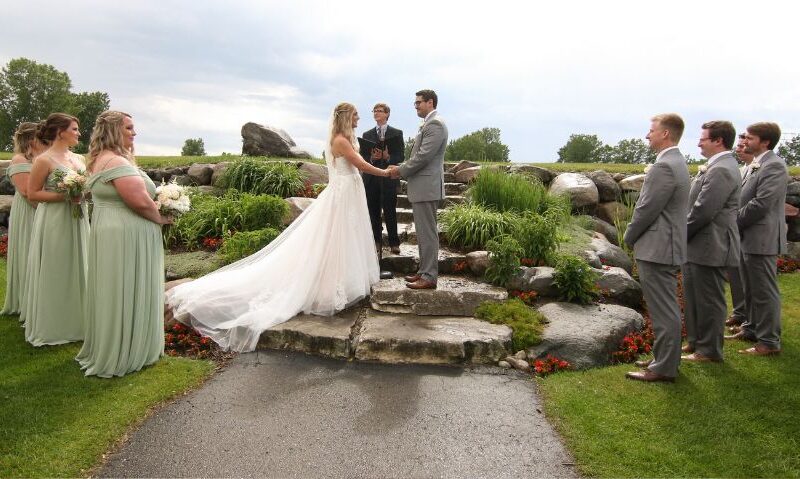Outdoor wedding ceremony with the bride and groom exchanging vows on a stone platform, surrounded by bridesmaids and groomsmen.
