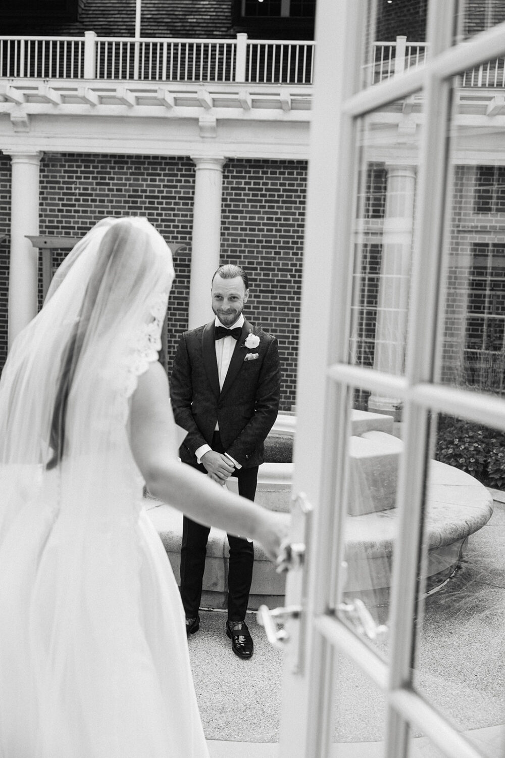 Groom smiling during a first look as the bride opens a door to reveal herself in a courtyard outside a brick building.