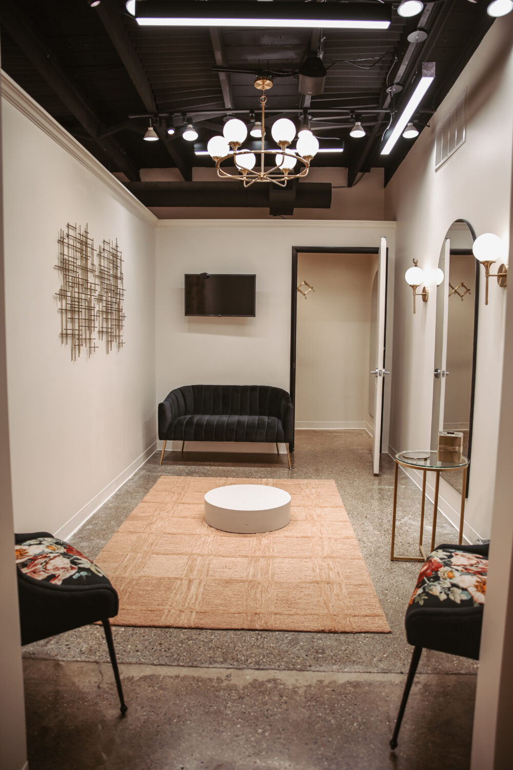 Modern bridal fitting suite with black velvet seating, arched mirrors, chandelier lighting, and a neutral area rug.