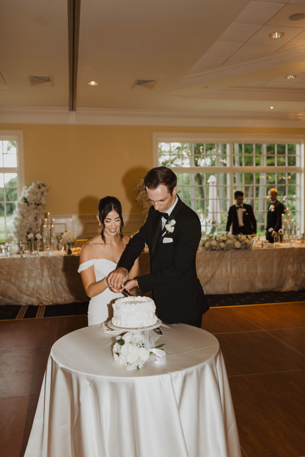 Bride and groom cut a white wedding cake together during their reception in an elegant ballroom with floral décor.
