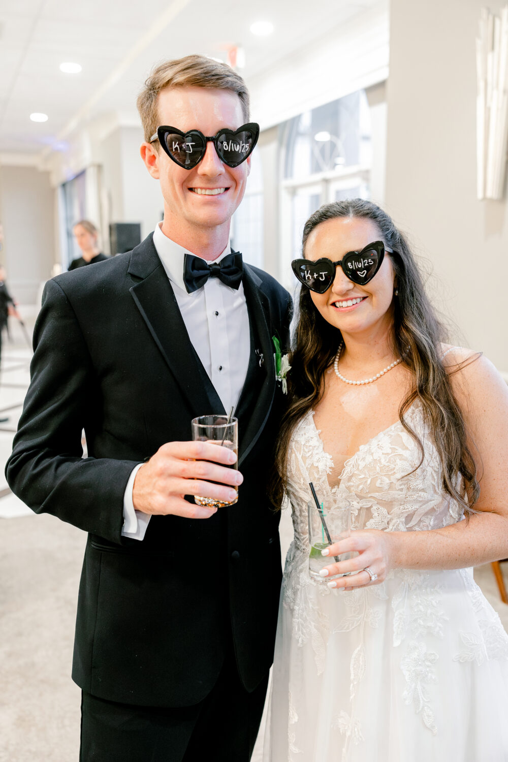 Bride and groom wearing heart-shaped sunglasses smile while holding drinks during their wedding reception.