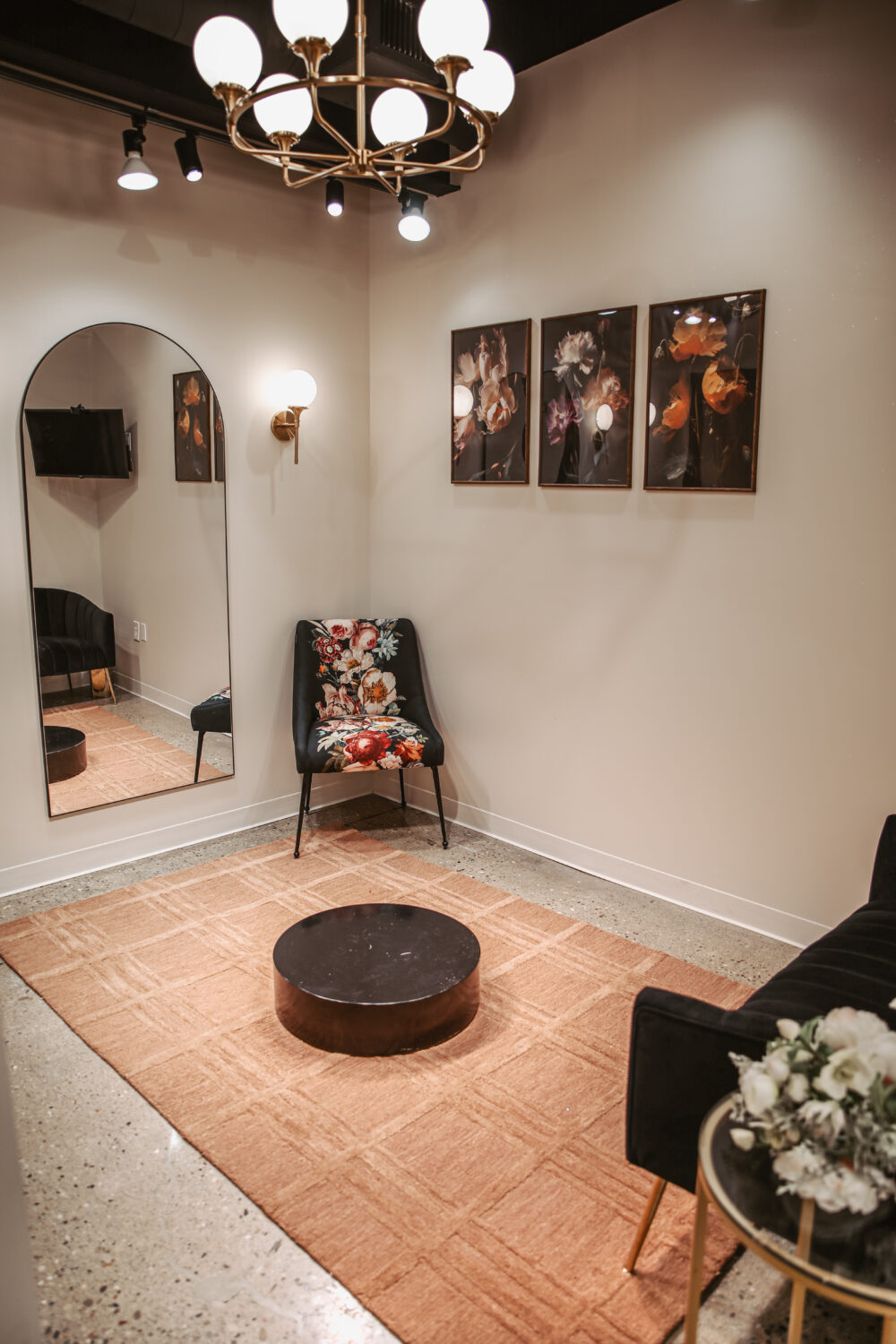 Elegant bridal appointment room featuring floral artwork, arched mirror, velvet seating, chandelier, and round coffee table.