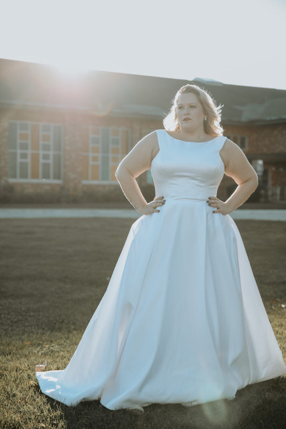 Bride wearing a sleeveless satin ball gown poses outdoors on a lawn with sunlight shining behind her.