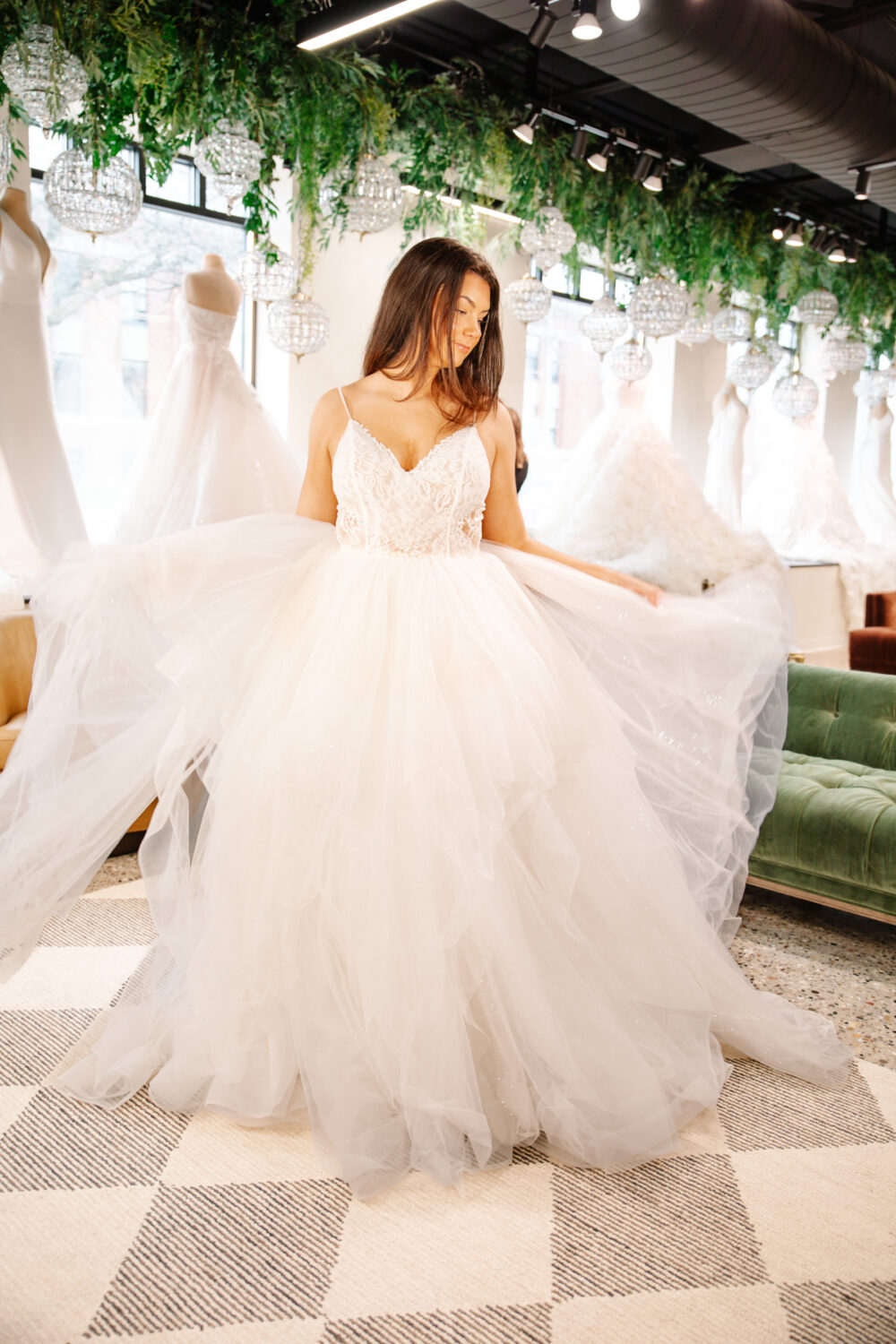 Bride in a voluminous tulle ball gown with a lace bodice holds out her skirt inside a bridal boutique filled with wedding dresses.