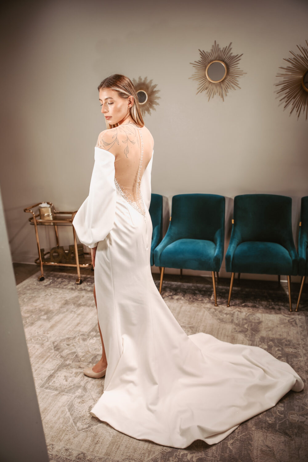 Woman modeling a fitted off-the-shoulder wedding gown with a low illusion back and long train in a bridal boutique.
