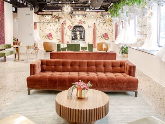 Spacious bridal boutique showroom with velvet seating, floral wall murals, chandeliers, and wedding gowns displayed along large windows.
