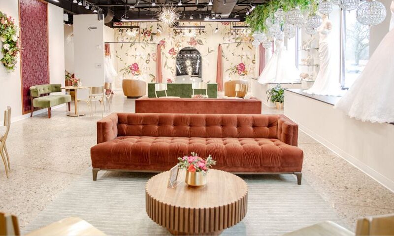 Spacious bridal boutique showroom with velvet seating, floral wall murals, chandeliers, and wedding gowns displayed along large windows.