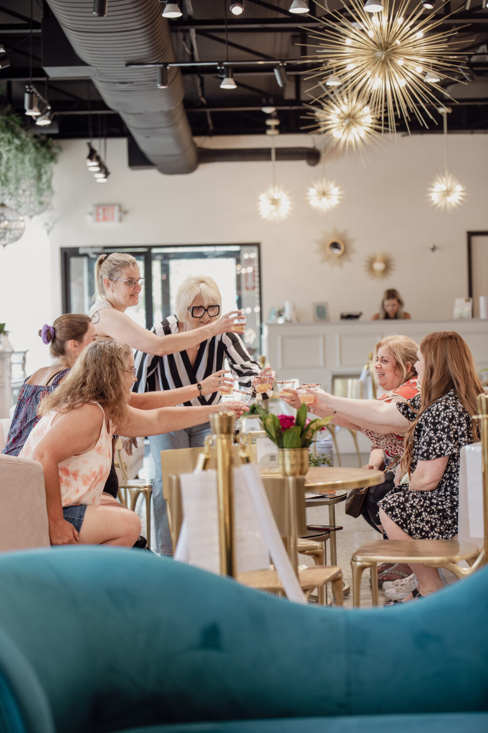 Group of women raising glasses in a bridal boutique lounge area during a celebration beneath modern chandeliers.