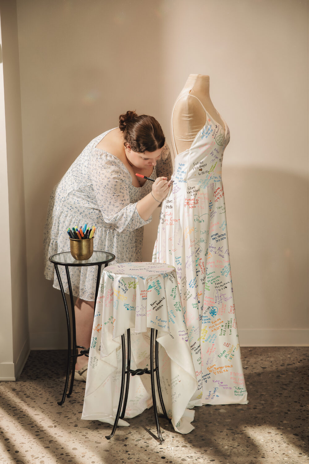 Woman signing messages on a white dress-covered display mannequin and matching tablecloth with colorful signatures and notes.