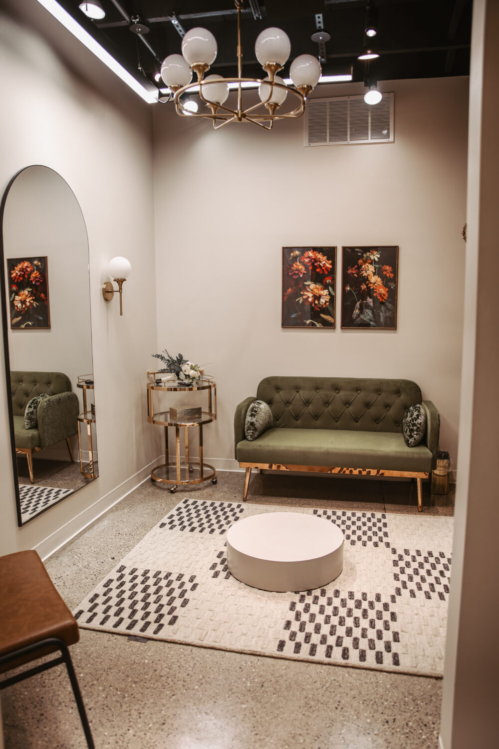 Private bridal fitting room with green tufted loveseat, large arched mirror, floral artwork, and modern chandelier lighting.