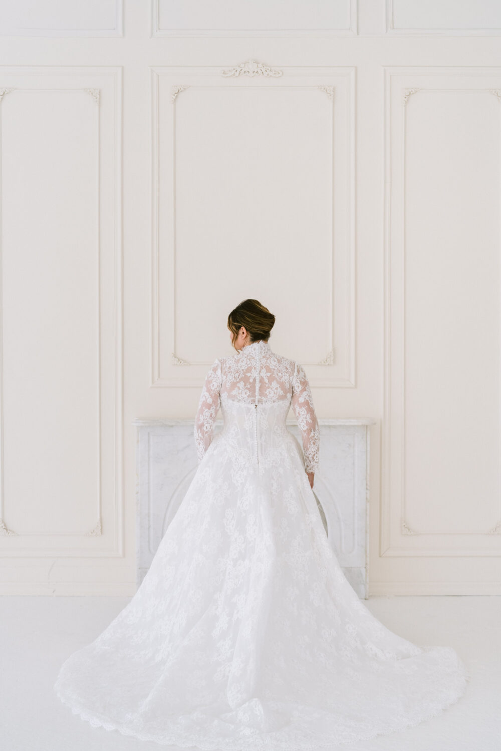 A bride stands with her back turned in a long-sleeve lace ball gown wedding dress with a flowing train in a white studio.