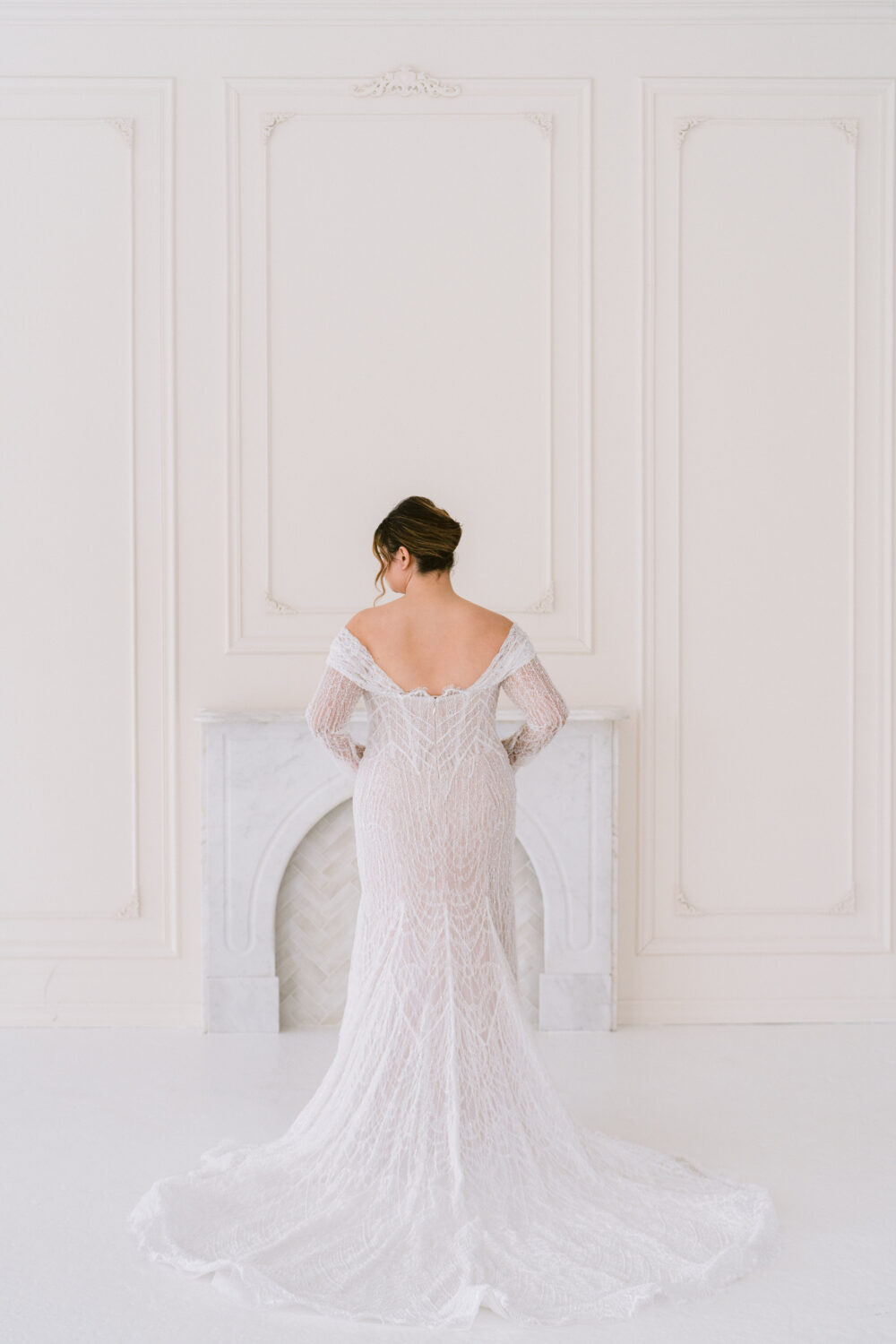 A bride models a fitted off-the-shoulder wedding dress with sheer sleeves and a long train while facing away from the camera in a white studio.