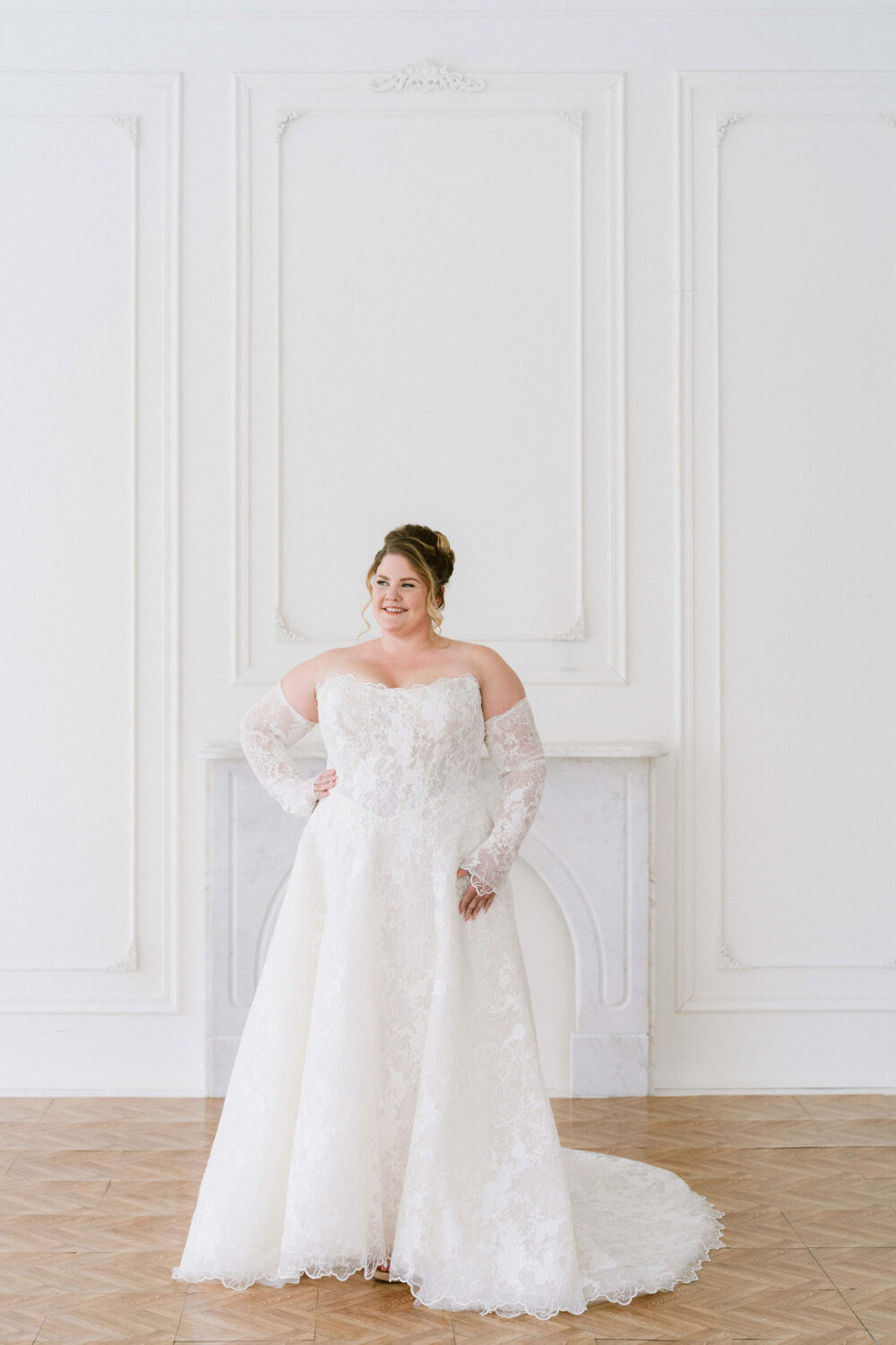 A smiling bride poses in an off-the-shoulder lace wedding gown with sheer sleeves and a long train in a bright white studio.