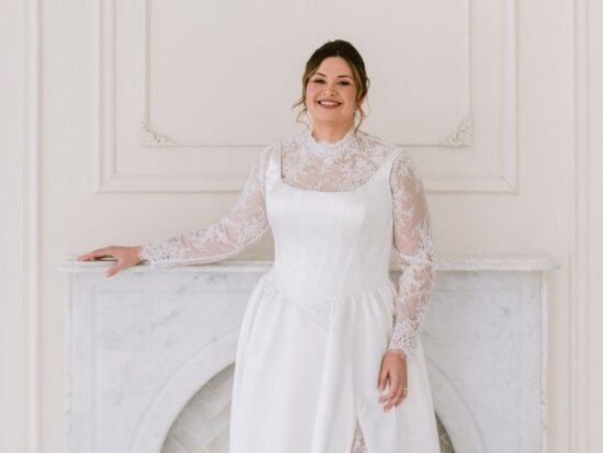 A smiling bride in a satin wedding gown with sheer lace sleeves stands in front of a marble fireplace in a bright white studio.