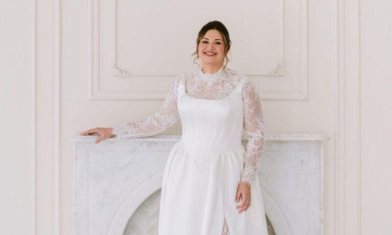 A smiling bride in a satin wedding gown with sheer lace sleeves stands in front of a marble fireplace in a bright white studio.