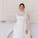 A smiling bride in a satin wedding gown with sheer lace sleeves stands in front of a marble fireplace in a bright white studio.