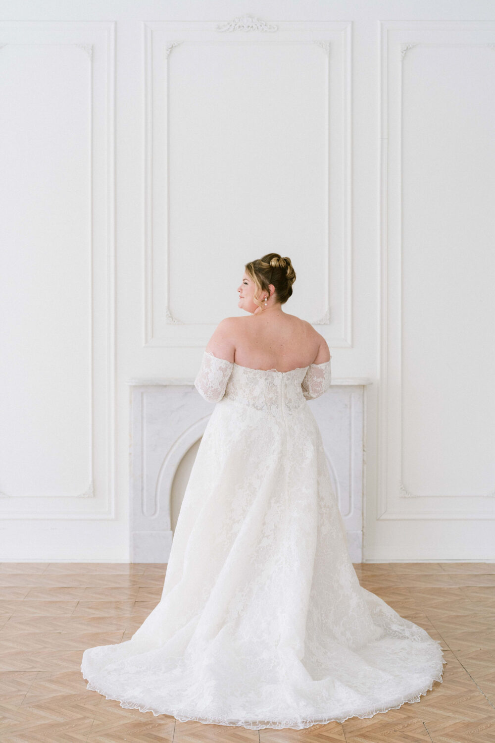 A bride wearing an off-the-shoulder lace wedding gown stands with her back turned in a bright white studio with decorative wall molding and a marble fireplace.