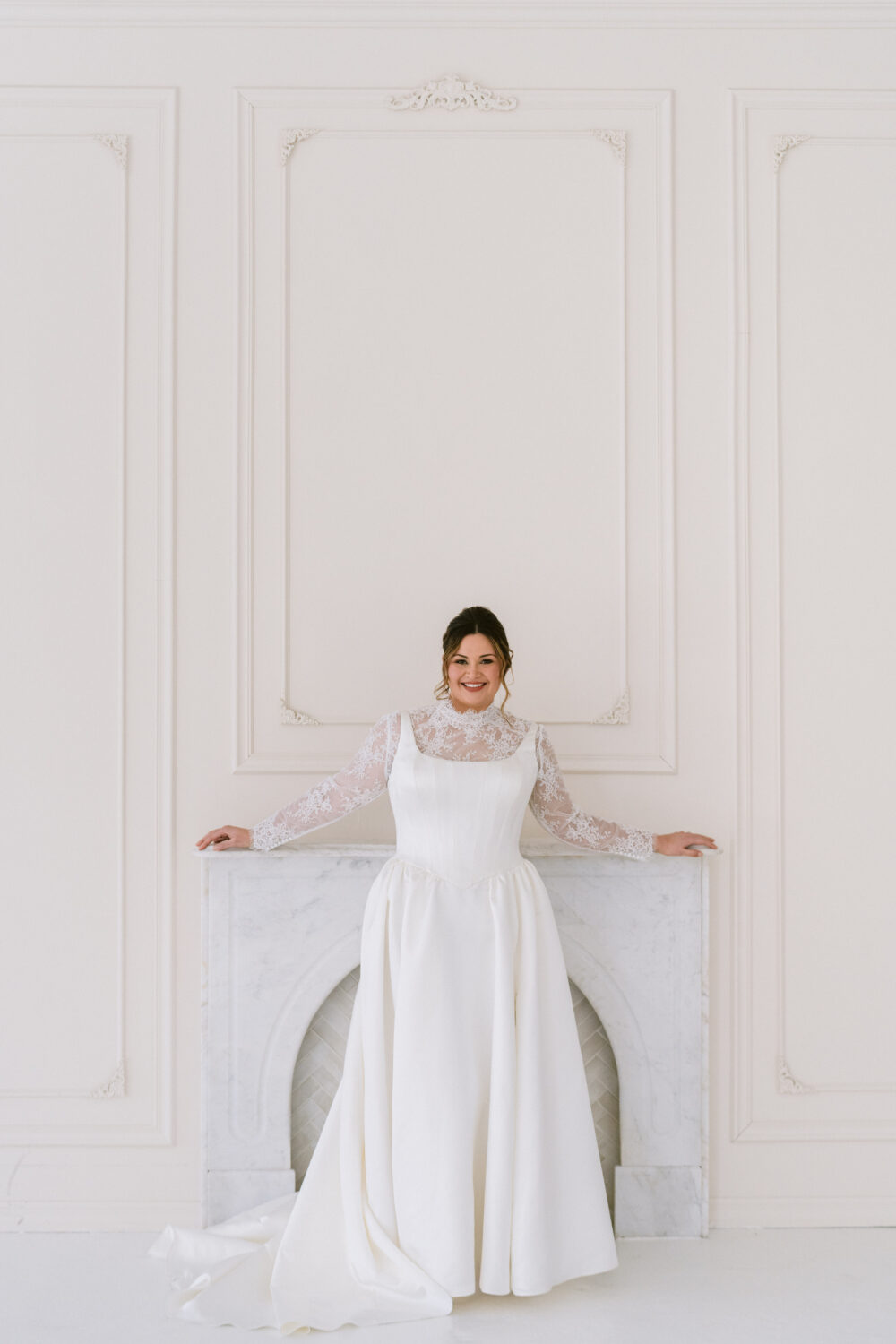 A smiling bride wears a long-sleeve lace wedding gown with a deep V neckline while standing in front of a white marble fireplace.