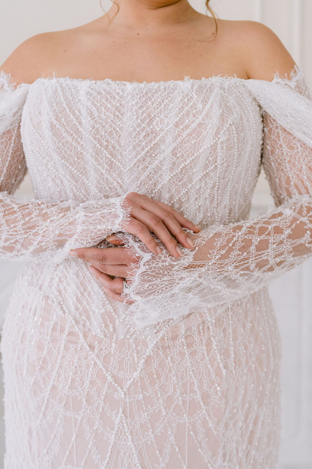 A close-up view of a fitted off-the-shoulder wedding dress with sheer lace sleeves, intricate beading, and delicate lace detailing across the bodice.