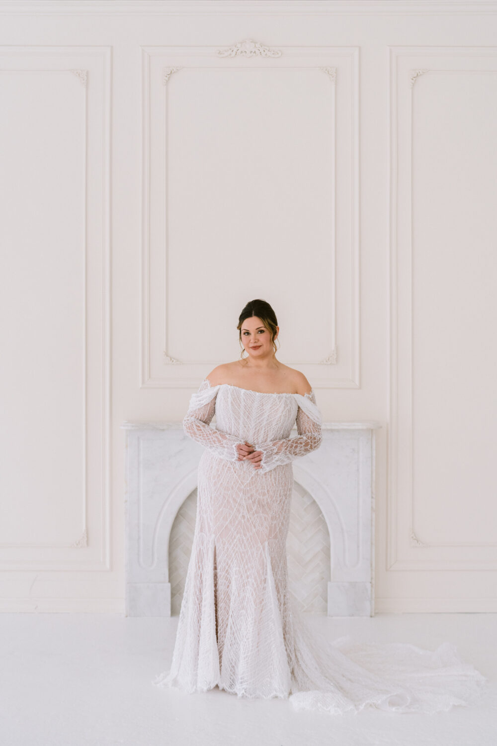 A bride poses in a fitted off-the-shoulder lace wedding gown with sheer sleeves and a long train in a bright white studio with a marble fireplace.