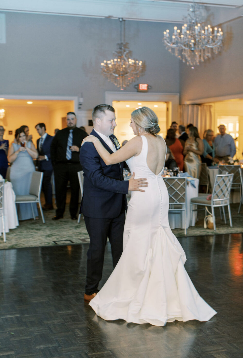 Bride and groom sharing their first dance in a reception ballroom beneath crystal chandeliers while guests watch.