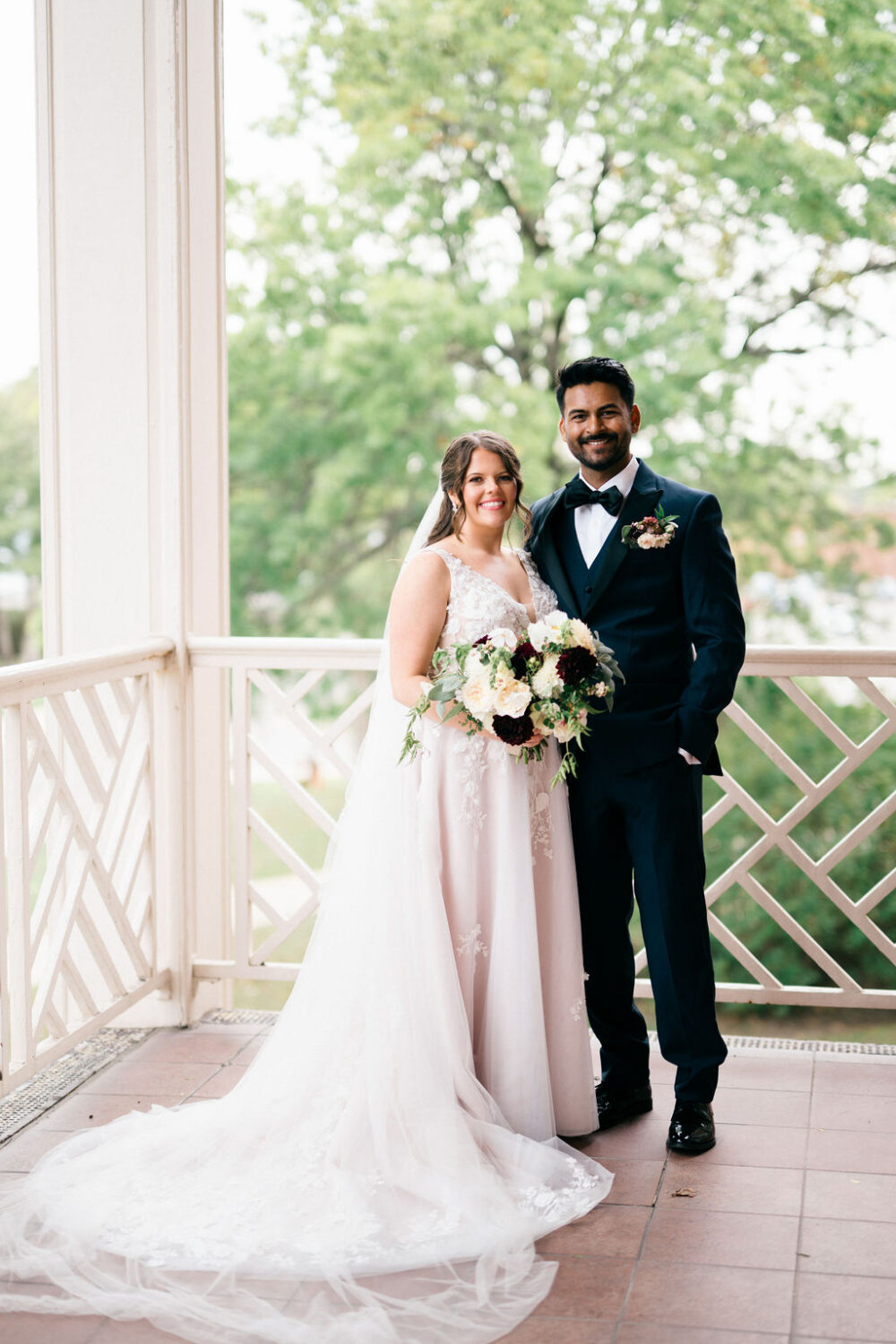 Bride and groom posing together on a covered porch, holding a bouquet and smiling at the camera.