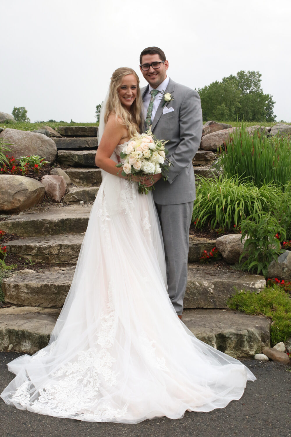 Bride and groom standing together in front of stone steps and landscaped gardens, holding a bouquet during an outdoor wedding portrait.