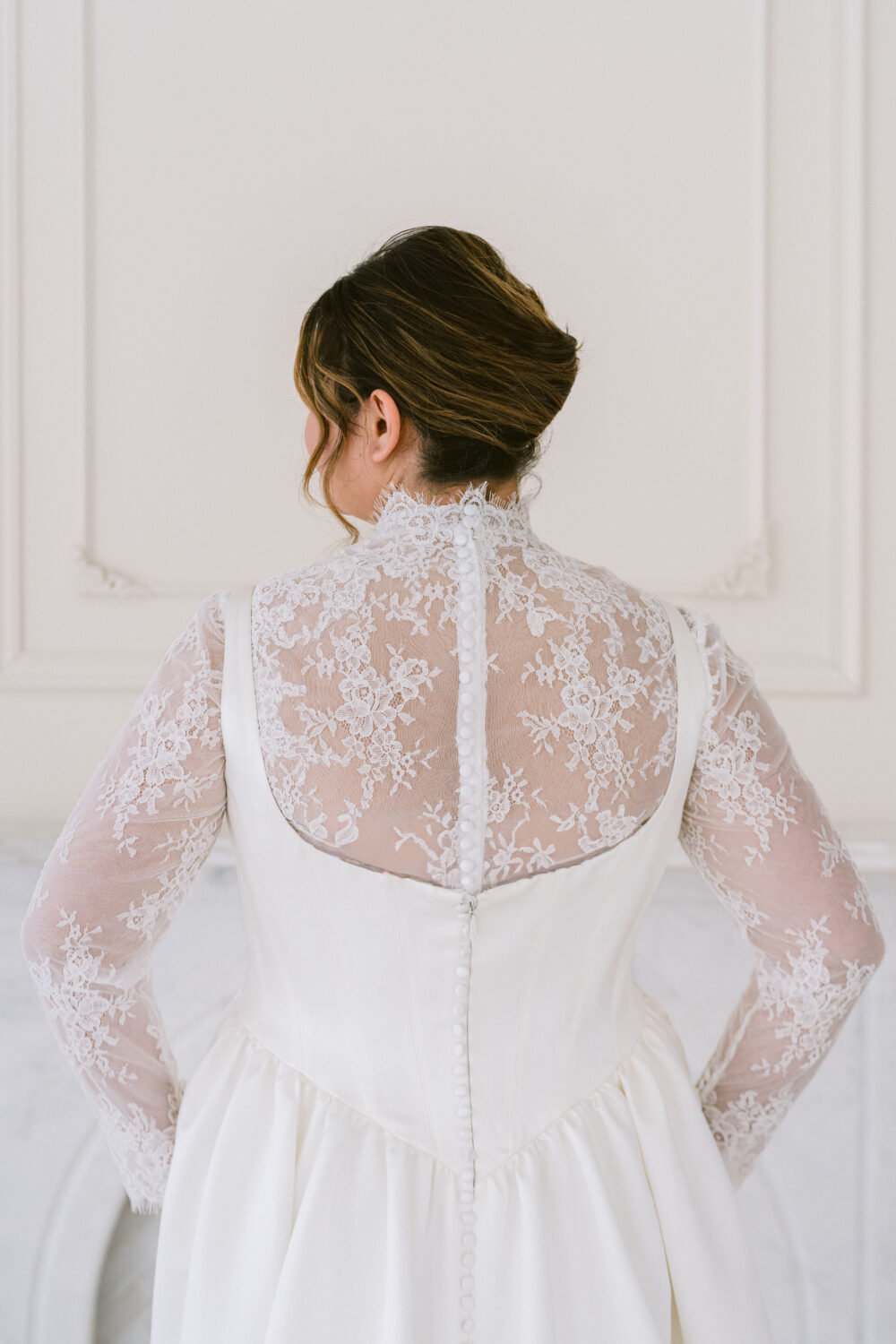 The back of a satin wedding gown with sheer lace long sleeves and button detailing is shown in a bright white studio.