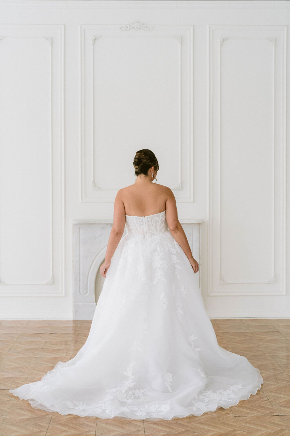 A bride stands with her back turned in a strapless floral appliqué wedding gown with a flowing train in a bright white studio.