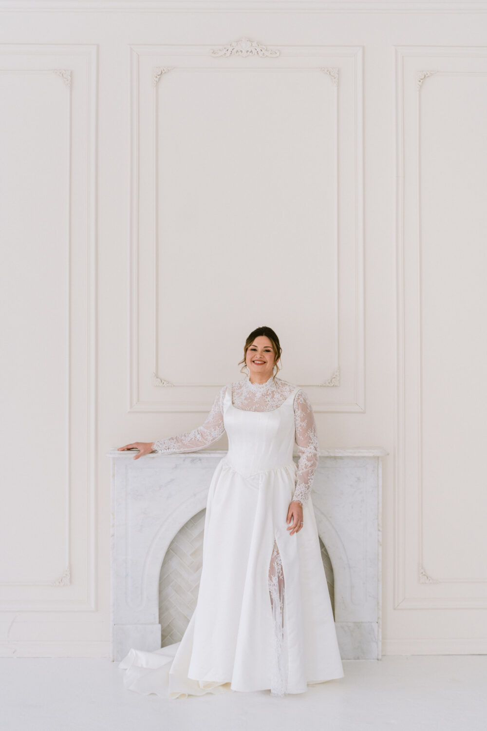 A smiling bride wears a satin wedding gown with sheer lace sleeves and a thigh-high slit while posing in a bright white studio.