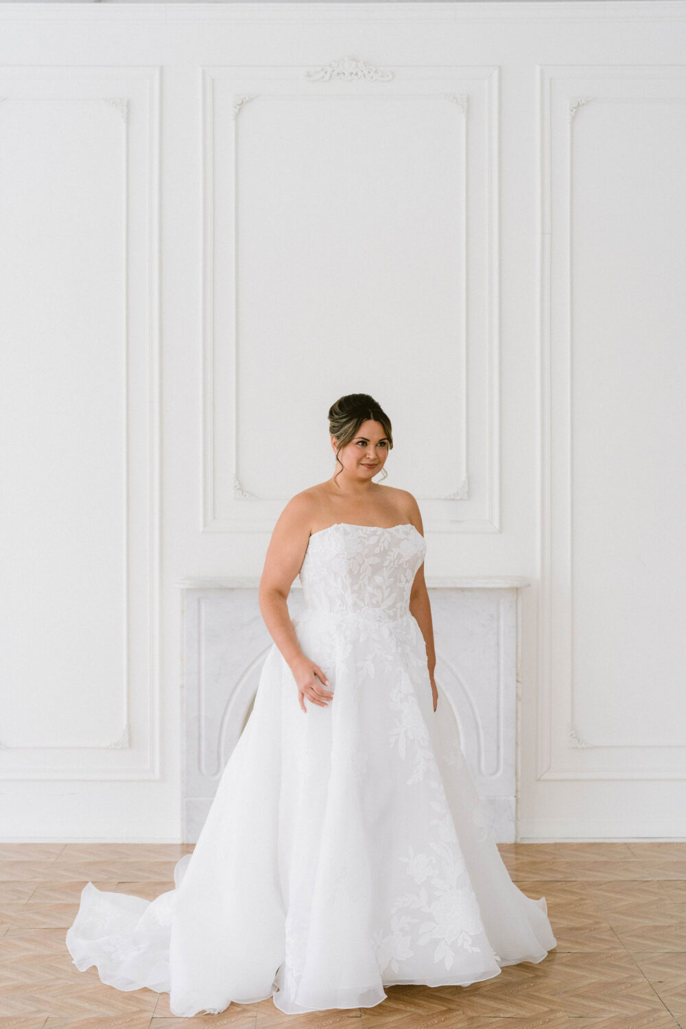 A bride poses in a strapless floral appliqué ball gown wedding dress in front of a marble fireplace in a white studio.