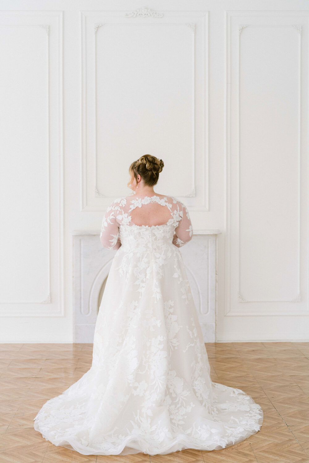 A bride stands with her back turned in a lace wedding gown with sheer sleeves and floral appliqué details in a white studio.