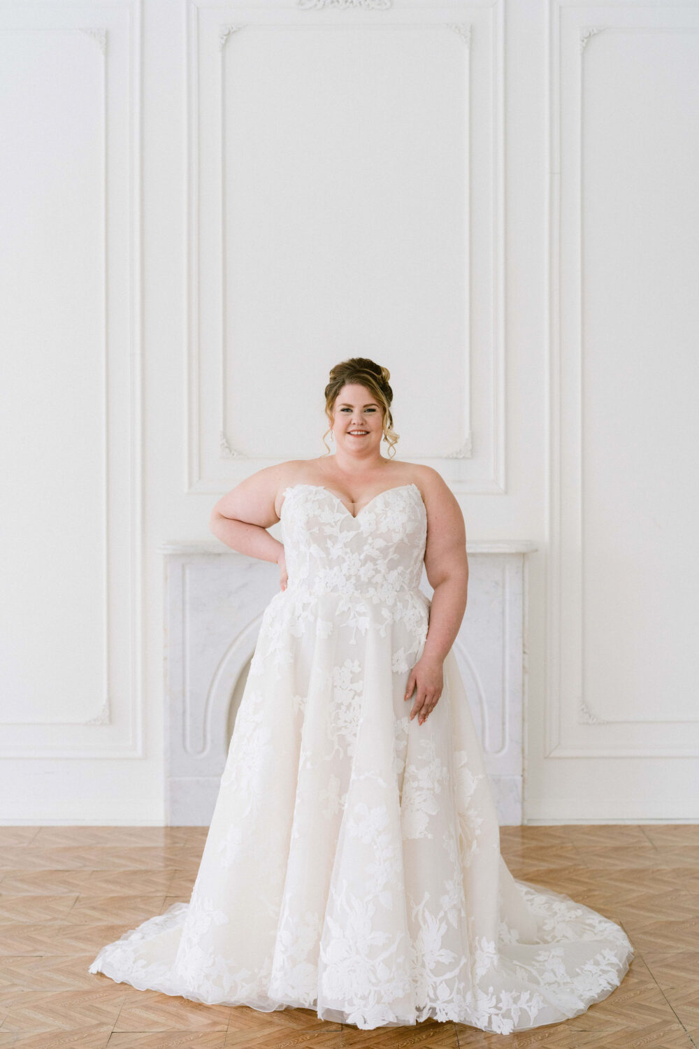 A smiling bride poses in a strapless floral appliqué wedding gown with a flowing train in front of a marble fireplace.